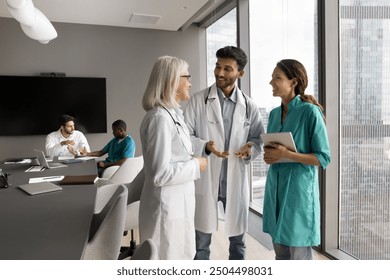 Three doctors, Indian, European and Hispanic internists in medical attire standing in modern clinic office, share opinion, skills, insights. Talk between experienced and young healthcare professionals - Powered by Shutterstock