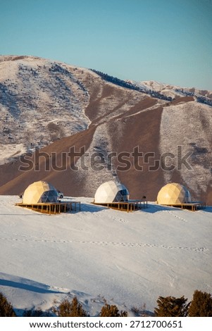 Three dome-shaped glamping accommodations situated on wooden platforms on a snow-covered slope against a background of arid, snowy mountains