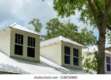 Three dormers with multiple double hung windows, a grey metal roof, and pale yellow vinyl siding on a large house. There are tall trees with thick green leaves both in front and behind the building. 