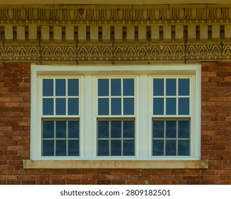 Three double-hung windows are set within a single unified white painted surround forming a triple window composition on the upper facade of a red brick commercial building in Abbeville, South Carolina. An elaborate cast stone or terra cotta cornice above the windows features multiple decorative bands including an egg-and-dart molding course, a row of stylized foliate motifs, and a dentil course at the top. The window panes reflect a deep blue sky. The image is captured in diffused ambient light with no direct shadows.