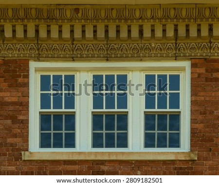 Three double-hung windows are set within a single unified white painted surround forming a triple window composition on the upper facade of a red brick commercial building in Abbeville, South Carolina. An elaborate cast stone or terra cotta cornice above the windows features multiple decorative bands including an egg-and-dart molding course, a row of stylized foliate motifs, and a dentil course at the top. The window panes reflect a deep blue sky. The image is captured in diffused ambient light with no direct shadows.