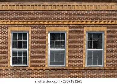 Three double-hung windows with white painted frames are set into the upper story of a red brick commercial building facade in Abbeville, South Carolina, each framed by contrasting tan brick surrounds forming decorative pilasters and lintels. Two horizontal decorative courses of tan brick run across the facade above and below the window row, creating a banded ornamental effect. The center window shows peeling paint along the wooden frame and deterioration of the glass surface. The image is captured in bright direct sunlight with strong contrast across the varied masonry surfaces.