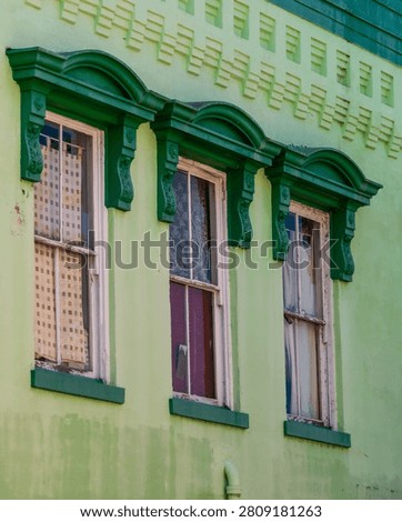 Three double-hung windows with white-painted frames showing deterioration and peeling paint are set into a light green stucco commercial building facade in Abbeville, South Carolina, each surmounted by a dark green ornate arched pediment with paired floral bracket corbels. A double-tier decorative cornice band with stepped geometric motifs runs horizontally across the upper facade. The lower section of the stucco wall shows areas of paint loss and surface staining. The image is captured from a slightly low angle in diffused ambient light.