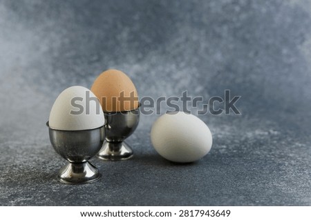 Three eggs two in silver holders and one on the surface are set against a gray background. The eggs have different colors and sizes and create a simple still life.