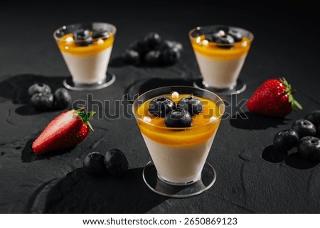 Three elegant dessert cups display creamy mousse topped with mango puree, blueberries, and strawberries on a dark surface. The composition emphasizes freshness and indulgence.