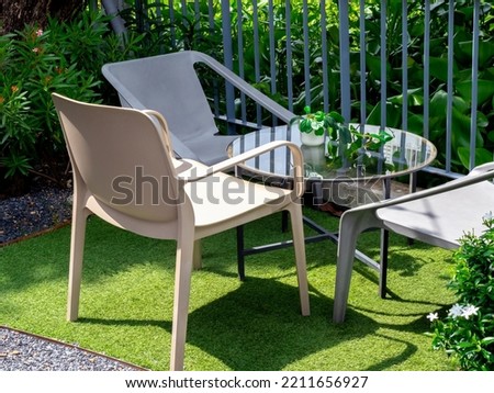 Three empty beige plastic chairs with round glass table with small plant pot in the outdoor garden on artificial grass floor and grey iron fence. Table set relaxing in garden.