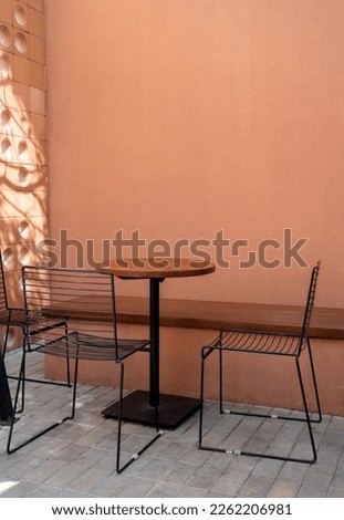 Three empty black wire chairs, long bench seat and round wooden table on gray brick blocks flooring and empty brown terracotta color wall background with copy space, vertical style.