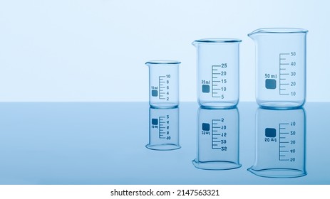 Three empty measuring beakers sitting on a mirror blue surface, glass lab containers standing in gradual order on a table, transparent light through laboratory flasks, precise comparing measurements  - Shutterstock ID 2147563321