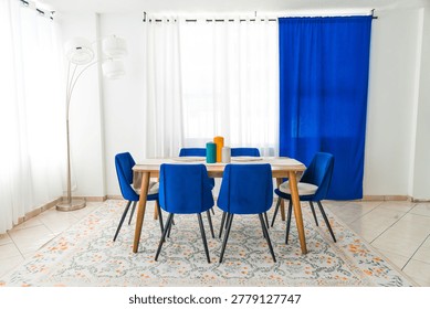 Three empty ribbed ceramic vases in teal, orange, and grey are placed in the center of a wooden dining table in a modern residential room. Blue velvet chairs are arranged around a table on a floral-patterned rug, set against a white wall, in a home dining area.