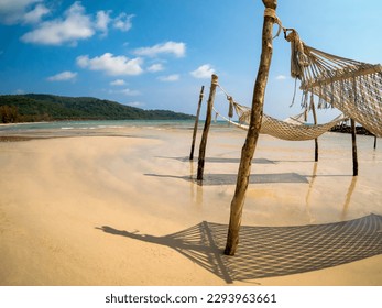 Three empty rope cradle hanging on the wooden poles on sandy beach, island and blue sky background, fisheye view. Beautiful shadow of hanging cradle or swing beds on clean sand beach.