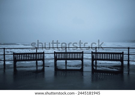 Three empty wooden benches on the seafront in Filey on the Yorkshire Coast in England. Taken at high tide on a moody, rainy day, with crashing waves and choppy sea on the beach below.