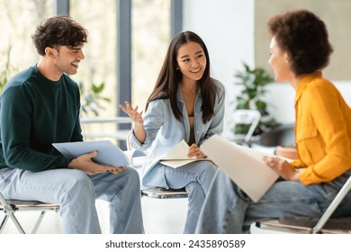 Three engaged diverse students sit in well-lit modern classroom, with one gesturing as they enthusiastically discuss the contents of their notebooks - Powered by Shutterstock