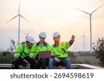 Three engineer in safety uniforms and helmets sitting on platform with wind turbines at sunset, discussing clean energy project. Concept of renewable energy, teamwork, sustainability and wind power.
