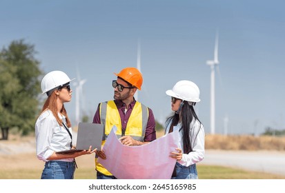 Three engineer are standing in a windmill field, looking at a blueprint. They are wearing hard hats and safety vests