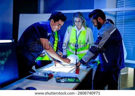 Three engineers leaning over blueprints on an illuminated planning table weighing a redesign option, a radio and hard hat within reach in the dark planning room indoors.