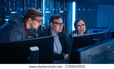 Three IT Engineers / Programmers Talking about Work, Using Computers. Technical Department with Data Server Racks. Software Development / Code Writing / Website Design / Database Architecture