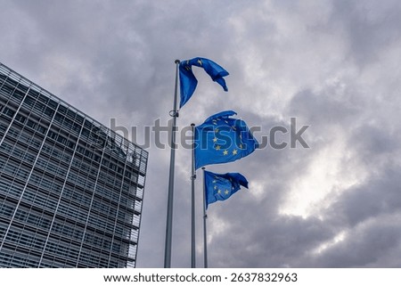 Three European flags wave dramatically against a dark, stormy sky