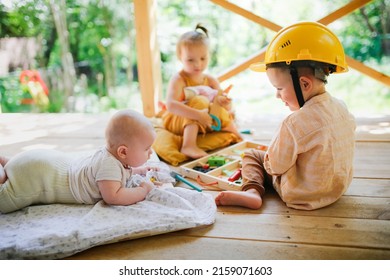 Three European sibling children play side by side on wooden porch. Newborn baby with older brother and sister. Summer in backyard, eco-friendly wooden safe toys