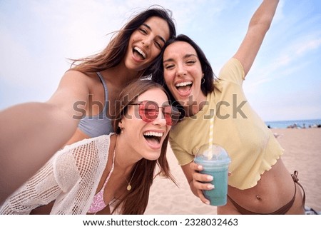 Three excited young Caucasian friends in summer clothes taking selfie on beach. Group of smiling women enjoying vacation. Cheerful beautiful girls of generation z pose for photo with mobile phone.