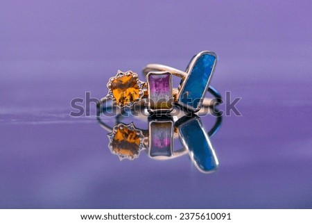 Three exquisite rings, one showcasing a radiant amber gem and the others featuring colorful rectangular stones, elegantly placed on a reflective lavender surface.
