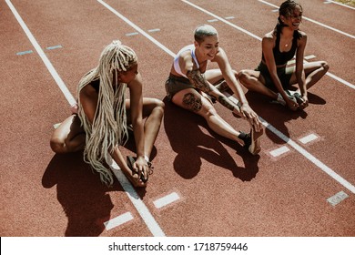 Three Female Athletes Sitting On A Running Track And Stretching Legs. Multi-ethnic Group Of Runners Doing Warmup Exercises At The Stadium.