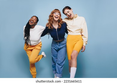 Three female friends laughing happily while embracing each other. Group of cheerful young women having fun while standing together against a blue studio background. - Powered by Shutterstock