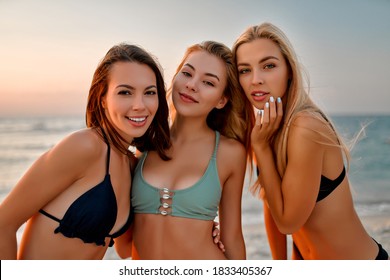 Three female friends in swimsuits having fun on the beach. Attractive girls near the sea smiling and looking at camera.