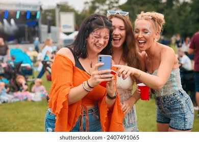 Three Female Friends Wearing Glitter Looking At Mobile Phone At Summer Music Festival Holding Drinks - Powered by Shutterstock