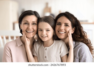 Three female generations family close up portrait. Adorable little 5s girl, granny 60s and young 35s mother sit on sofa, look at camera, enjoy time together, feeling love, having strong family ties