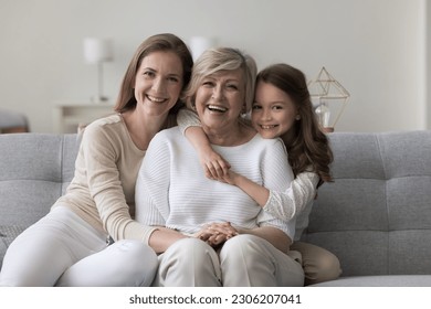 Three female generations family portrait. Cute little girl hugging tightly her beloved granny sit on sofa with young mom, smile look at camera, enjoy leisure time together. Unconditional love and ties - Powered by Shutterstock