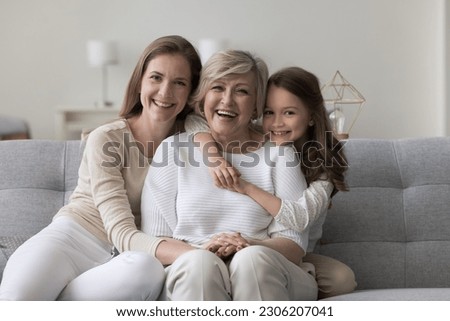 Three female generations family portrait. Cute little girl hugging tightly her beloved granny sit on sofa with young mom, smile look at camera, enjoy leisure time together. Unconditional love and ties