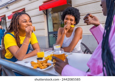 Three female young african friends eating fast food with beer in an outdoor restaurant - Powered by Shutterstock