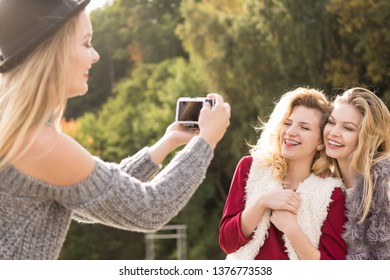 Three females friends having fun during outdoor photo session. Woman taking pictures of two during warm autumn weather.