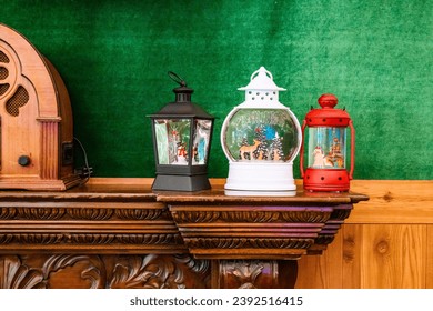 Three festive lanterns and a snow globe with Christmas scenes on a carved wooden mantelpiece against a green wal - Powered by Shutterstock