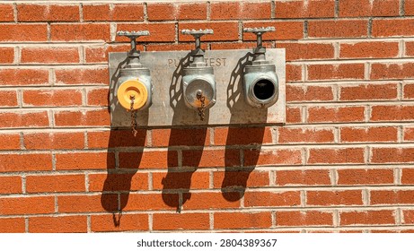 Three fire hydrants mounted on a brick wall show connections for testing. One hydrant has a yellow cap, while the other two are grey and black.