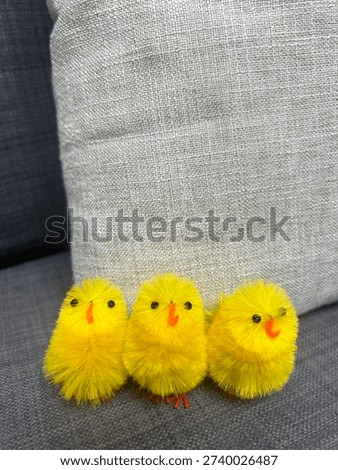 Three fluffy yellow chick decorations sitting on a gray fabric couch with a neutral pillow in the background, creating a cute spring and Easter holiday concept with copy space.