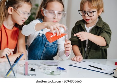 Three Focused Little Kids Doing Home Science Project, Carefully Filling A Flask From The Beaker. All Behid Table, Wearing Glasses. Chemical Glassware And Colored Liquids On The Table.