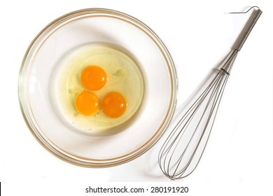 Three fresh raw eggs in a glass bowl and whisk for whipping on a white background. Top view