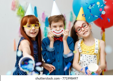 Three friendly children in festive cone caps and big eyewear, stand in decorative room with balloons, have fun together as celebrate birthday look with happy expressions at camera, enjoy playing games