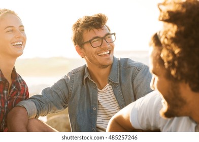 Three Friends Enjoying Together At Sunset. Young Woman And Smiling Man With Spectacles Having Fun With Friends. Happy Guys And Girl In Casual Sitting Outdoor.