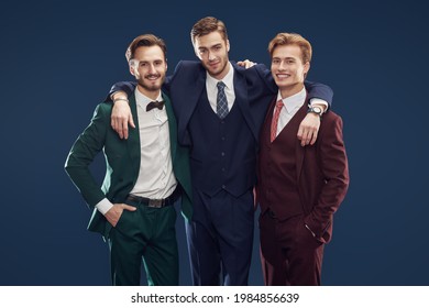 Three Friends. Men's Fashion. Three Handsome Men In Elegant Suits Hug Each Other In A Friendly Manner And Smile. Dark Blue Background.