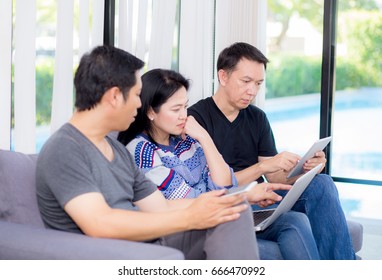 Three Friends On Line With Multiple Devices And Talking Sitting On A Sofa In The Living Room In A House Interior.