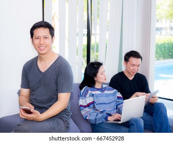 Three Friends On Line With Multiple Devices And Talking Sitting On A Sofa In The Living Room In A House Interior.