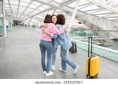 Three friends share a heartfelt hug at a bustling airport terminal. One is wearing a pink sweater, and the others are in a light blue outfit, with a yellow suitcase nearby, ready for their journey. - Powered by Shutterstock