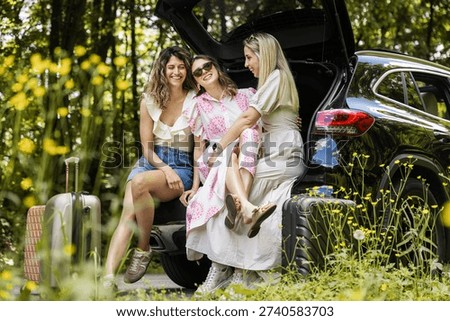 Three friends sit in the open trunk of a car parked in a green field. They smile and laugh while enjoying each other's company. Their luggage rests nearby on the ground.