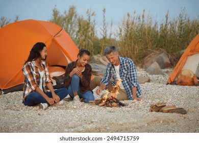 Three friends sitting by a campfire at their beachside campsite with orange tents, sharing stories and enjoying the evening together. - Powered by Shutterstock