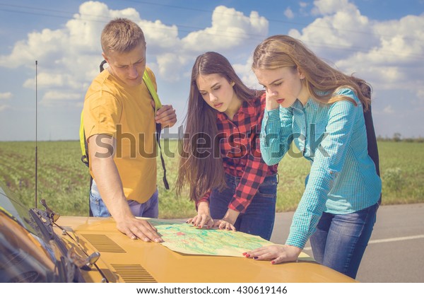 Three friends wanderer\
searching direction on location map on old car, two girls and  guy\
tourist searching road to hotel or direction seaon atlas in country\
road