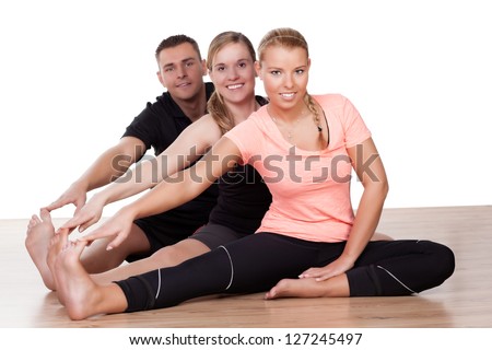 Three friends working out in a gym sitting on the wooden floor doing stretching exercises with their legs extended