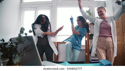 Three Fun Happy Multiethnic Female Doctors Dance Together At Work Lab Celebrating Victory Over Coronavirus Pandemic.