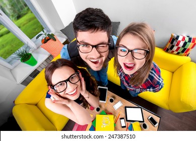 Three funny nerds looking together at camera standing in the room with couch and different digital stuff on background